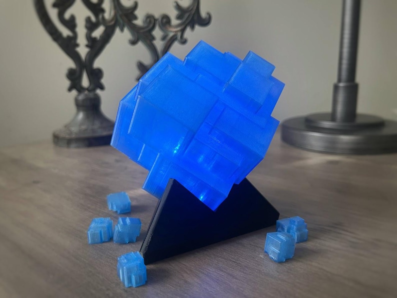 Puede incluir: Un objeto azul en forma de cubo impreso en 3D sobre un soporte triangular negro. El cubo est&aacute; iluminado desde el interior, lo que le da un brillo azul. Hay varios cubos azules m&aacute;s peque&ntilde;os dispersos alrededor de la base.