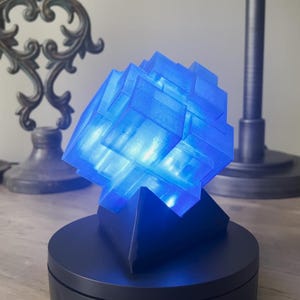 May include: A blue light-up cube lamp with a geometric design. The lamp is on a black base and is sitting on a wooden table.