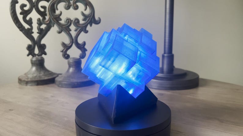 Puede incluir: Una l&aacute;mpara de cubo azul iluminada con un dise&ntilde;o geom&eacute;trico. La l&aacute;mpara est&aacute; sobre una base negra y est&aacute; sentada sobre una mesa de madera.