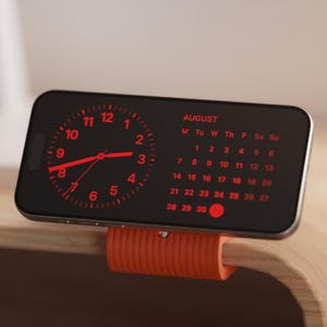 Può includere: Uno smartphone nero mostra un orologio rosso e un calendario di agosto. Il telefono è montato su un supporto cilindrico arancione, appoggiato su una superficie di legno. L'orologio mostra l'ora e il calendario evidenzia la data.