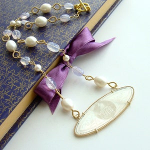 May include: A gold necklace with a large oval pendant featuring an intricate design. The necklace is made with white pearls and light pink stones.
