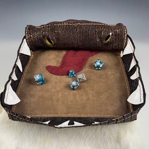 May include: A brown leather dice tray with a crocodile pattern and a red tongue. The tray has white teeth around the edge and is filled with blue and gold dice.