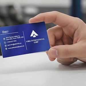 May include: A blue business card held in a hand. The card displays the text "Henry & Betty Co." and "YORK INTERNATIONAL CORP." alongside contact details. The background is blurred, with a white surface in the foreground.