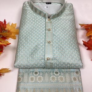 May include: A light blue kurta pajama set with a white and gold patterned design. The kurta has a mandarin collar and a button closure. The pajama pants have a floral design.