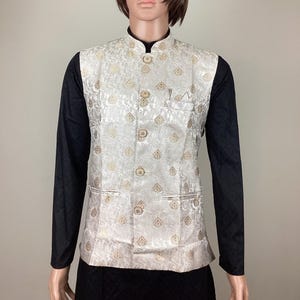May include: A white brocade waistcoat with gold leaf pattern and button closures. The waistcoat has a pocket on the left side.