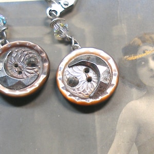 Puede incluir: Un par de pendientes con botones de estilo antiguo con un diseño de media luna. Los botones están hechos de un material marrón y plateado y tienen una superficie texturizada. Los pendientes están unidos a cadenas plateadas con cuentas de cristal.