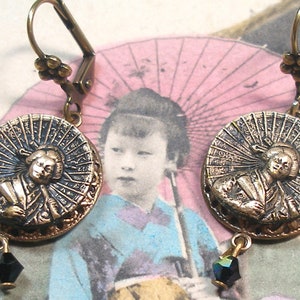 May include: A pair of gold-tone earrings with a black glass bead dangle. Each earring features a round, ornate, metal charm with a raised image of a woman holding a fan.