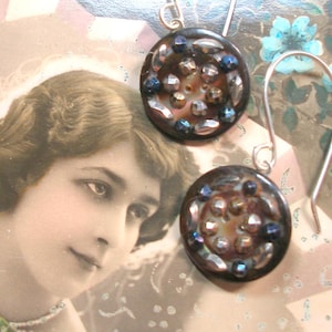 May include: A pair of earrings with a dark brown base and iridescent blue and silver accents. The earrings are shaped like buttons with a circular design.