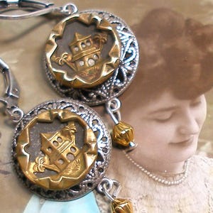 Może przedstawiać: Zbliżenie pary srebrnych kolczyków ze złotymi i srebrnymi akcentami. Każdy kolczyk ma okrągły wzór ze złotym budynkiem pośrodku, otoczonym srebrną filigranową obwódką i małą złotą koralikiem.