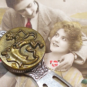 May include: A gold-toned bookmark with a round, embossed design featuring a lion. The lion is depicted standing on its hind legs with its head turned to the side. The bookmark is attached to a silver-toned metal handle.