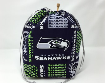 Puede incluir: Una bolsa de cordón azul con un logotipo de los Seattle Seahawks y las palabras "Seattle Seahawks" repetidas en verde y blanco. La bolsa tiene un cordón blanco y un botón de madera.