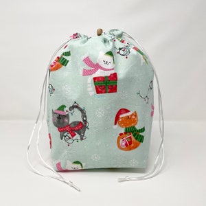 May include: A light blue drawstring bag with a Christmas cat print. The bag features cats wearing Santa hats and holding presents, as well as cats wearing scarves and hats with snowflakes and Christmas lights in the background.