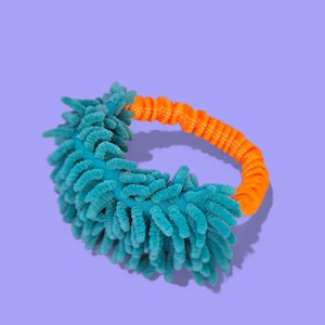 May include: A teal microfiber duster with an orange elastic handle. The duster has a curved shape and is designed for cleaning. The background is a solid purple color.