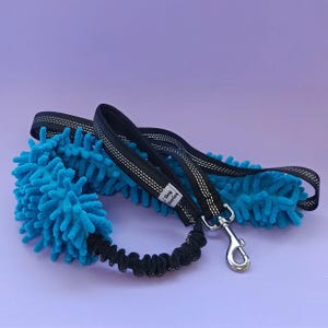 Op de afbeelding: Een zwarte hondenriem met een blauwe microvezel reinigingsaccessoire. De riem heeft een zilveren clip en een zwart bungee-koordgedeelte. Het label op de riem luidt "The Dog Diaries".