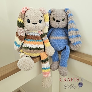 Peut inclure: Deux peluches de lapin crochetées à la main. L'un est beige avec des oreilles et des membres rayés colorés. L'autre est gris et bleu. Les deux ont des yeux en bouton et sont assis sur une étagère en bois. Le texte "Crafts by Linda" est en bas à droite.