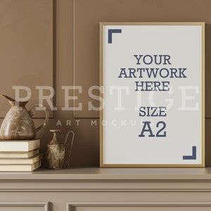 Op de afbeelding: Een gouden geframed poster mockup met de tekst "YOUR ARTWORK HERE SIZE A2" op een bruine muur met een plank en decoratieve items.