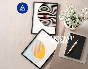 May include: Two framed art prints with black frames. One print features a black and white striped design with a red eye. The other print has a yellow and orange geometric design. A notebook, pen, and flowers are also in the image.