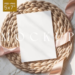 May include: A 5x7 inch white card mockup with the word "MOCKUP" in large white letters, resting on a woven beige placemat. A blush pink ribbon is draped around the card and placemat. The background is a marble surface.