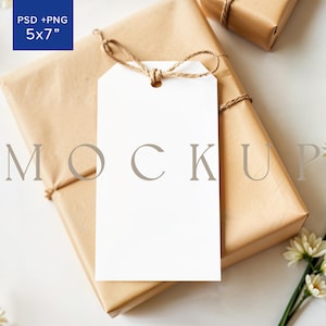 May include: A white gift tag with a brown twine bow tied around it. The tag is sitting on top of a brown gift box with the text "MOCKUP" printed on it. The gift box is wrapped in brown paper and tied with twine. There are white flowers in the background.