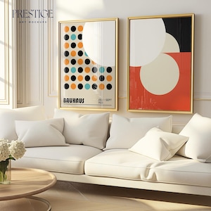 Op de afbeelding: Twee ingelijste abstracte kunstprints met geometrische vormen in zwart, wit, rood, oranje en blauw. De prints hangen aan een witte muur boven een witte bank met witte kussens.