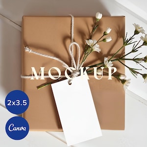 Puede incluir: Una caja de regalo cuadrada marrón atada con hilo y una etiqueta blanca en blanco. Flores blancas y tallos verdes están metidos debajo del hilo. La palabra "MOCKUP" está impresa en la caja. Un círculo azul con el texto "2x3.5" y otro con "Canva" están en la esquina inferior izquierda.