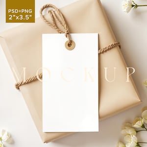 Puede incluir: Una etiqueta de regalo blanca en blanco con un anillo de metal dorado unido a una cuerda de yute marrón. La etiqueta está atada a una caja de regalo marrón envuelta en papel marrón. Flores blancas están esparcidas alrededor de la caja.