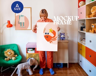Può includere: Una persona tiene in mano un grande poster bianco con un'illustrazione di un granchio dei cartoni animati. Il granchio è arancione con occhi blu e una stella gialla. Il poster si trova in una stanza con una sedia verde, peluche e un mobile contenitore colorato. Il testo "MOCKUP FRAME" è visibile.