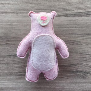 May include: A handmade pink bear plush toy with a light gray belly. The bear has small black eyes, a pink nose, and a white and pink face. The toy is made of soft fabric and has stitched details.