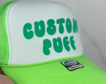 May include: A white and green trucker hat with the text "CUSTOM PUFF" in green letters. The hat has a mesh back and a green brim.