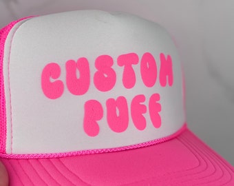 May include: A white trucker hat with a pink mesh back and a pink "CUSTOM PUFF" logo.