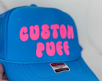 May include: A blue trucker hat with a pink "Custom Puff" logo. The hat has a mesh back and a foam front.