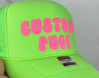 May include: A bright green trucker hat with a pink "CUSTOM PUG" logo. The hat has a mesh back and a black "OTTO WORLD FAMOUS FOAM FRONT CAPS" label.