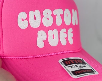 May include: A pink trucker hat with a white foam front and the text "CUSTOM PUFF" in white bubble letters. The hat has a black and white label that says "OTTO WORLD FAMOUS FOAM FRONT OSFM TRUCKER DURABLE SNAP CLOSURE".