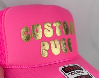 May include: Pink trucker hat with a gold "Custom Puff" logo. The hat has a mesh back and a snap closure.