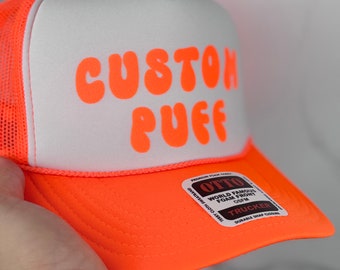 May include: Orange and white trucker hat with the text "CUSTOM PUFF" in orange bubble letters. The hat has a mesh back and a foam front.