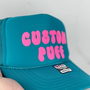 May include: A teal blue trucker hat with a pink "CUSTOM PUFF" logo. The hat has a mesh back and a black "OTTO WORLD FAMOUS FOUR FRONT" label.