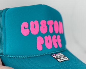 May include: A teal blue trucker hat with a pink "CUSTOM PUFF" logo. The hat has a mesh back and a black "OTTO WORLD FAMOUS FOUR FRONT" label.