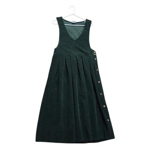 May include: A dark forest green corduroy pinafore dress featuring a V-neckline, a sleeveless bodice, and a pleated skirt. The side of the dress is secured with a vertical row of small, round, light-colored buttons extending from the waist down.