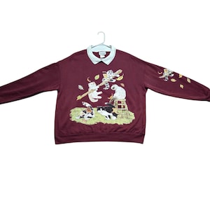 May include: A maroon long-sleeve sweatshirt with a white collared neckline. The front features a graphic of multiple cats playing on a branch and a stool amidst falling leaves. A smaller cat graphic is printed on the left sleeve.