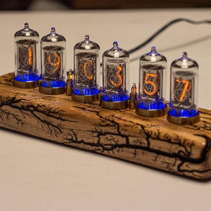 May include: A digital clock with six glass tubes displaying orange numbers, mounted on a wooden base with a unique, patterned grain. Each tube has a blue light at the base. The clock is a unique piece of home decor.