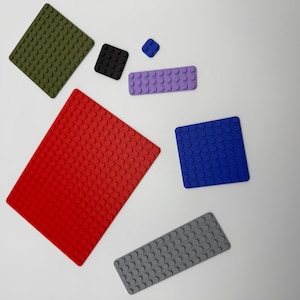 Peut inclure: Un assortiment de tapis et dessous de verre en silicone texturé, de style blocs de construction. Les pièces sont de différentes formes et tailles, notamment rouges, bleues, vertes, noires, violettes et grises.