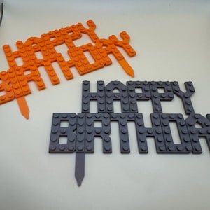 3D printed orange and dark gray building block style "Happy Birthday" cake toppers for kids' parties.