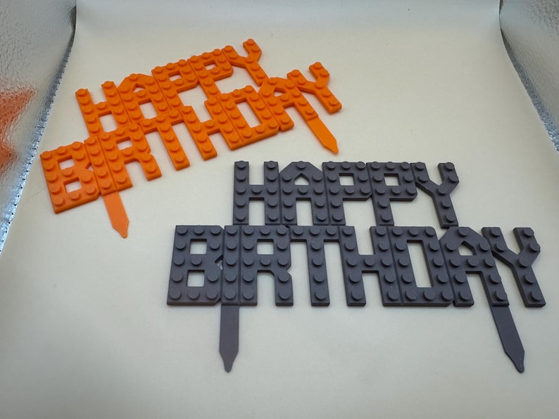 3D printed orange and dark gray building block style "Happy Birthday" cake toppers for kids' parties.