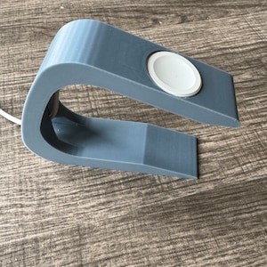 Könnte beinhalten: Ein grauer 3D-gedruckter Ständer zum Aufladen einer Smartwatch. Der Ständer hat einen kreisförmigen Ausschnitt für das Zifferblatt der Uhr und eine gebogene Basis.
