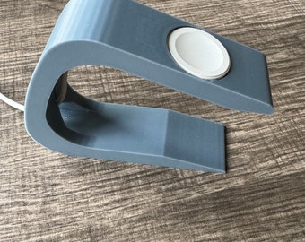 Op de afbeelding: Een grijze 3D-geprinte standaard voor het opladen van een smartwatch. De standaard heeft een ronde uitsparing voor het horloge en een gebogen basis.