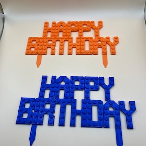 Two 3D printed "Happy Birthday" cake toppers in orange and blue, featuring a textured building block or brick-style design.