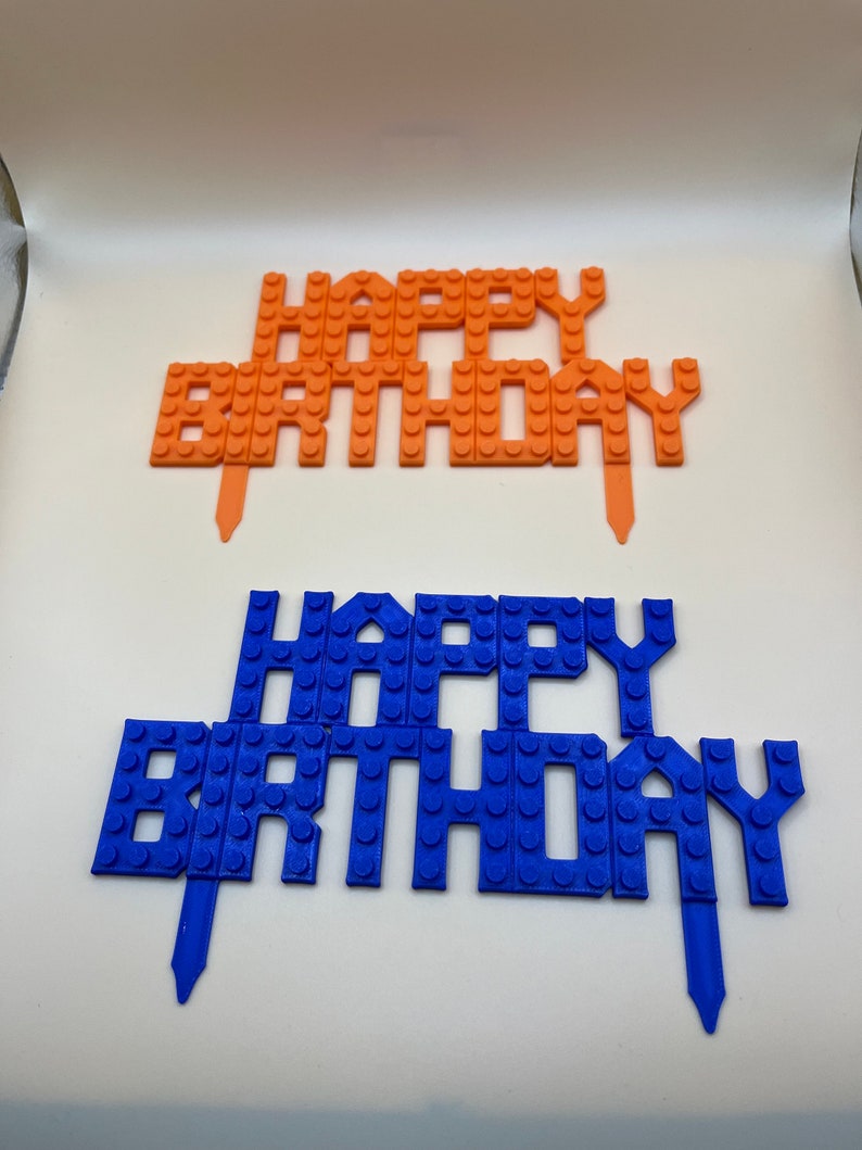 Two 3D printed "Happy Birthday" cake toppers in orange and blue, featuring a textured building block or brick-style design.