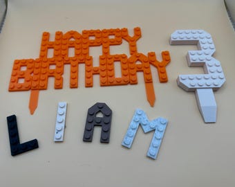 Op de afbeelding: Oranje en witte Lego-achtige taarttoppers die "Happy Birthday" en "Liam" spellen met een witte nummer 3 topper.