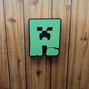 May include: A green and black Minecraft Creeper face decoration. The Creeper face is a square shape with a green background and black details. The decoration is mounted on a wooden surface.