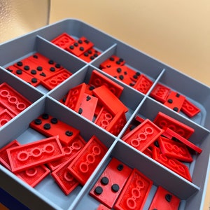 Peut inclure: Une boîte de rangement en plastique gris avec des compartiments remplis de dominos rouges. Les dominos ont des points noirs.